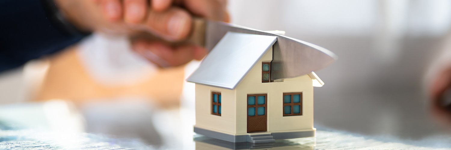 Twee handen die samen met een mes een klein modelhuisje doormidden snijden ter illustratie van de verdeling van een woning bij echtscheiding en huwelijksvermogensrecht.
